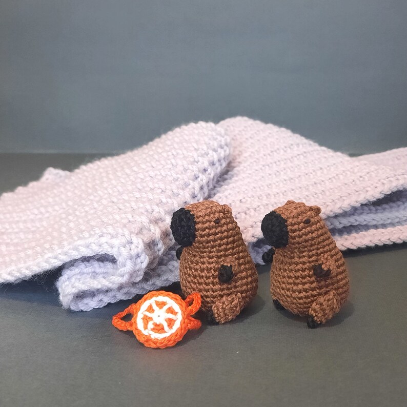 May include: Two brown crocheted animal figurines with black eyes and noses, sitting on a light purple knitted blanket. A small orange crocheted circle with a white centre is in the foreground.