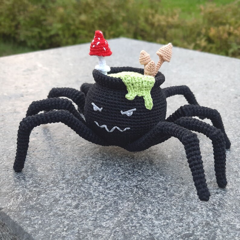 May include: A black crocheted spider with eight legs carrying a cauldron with green slime and a red and white mushroom on top.