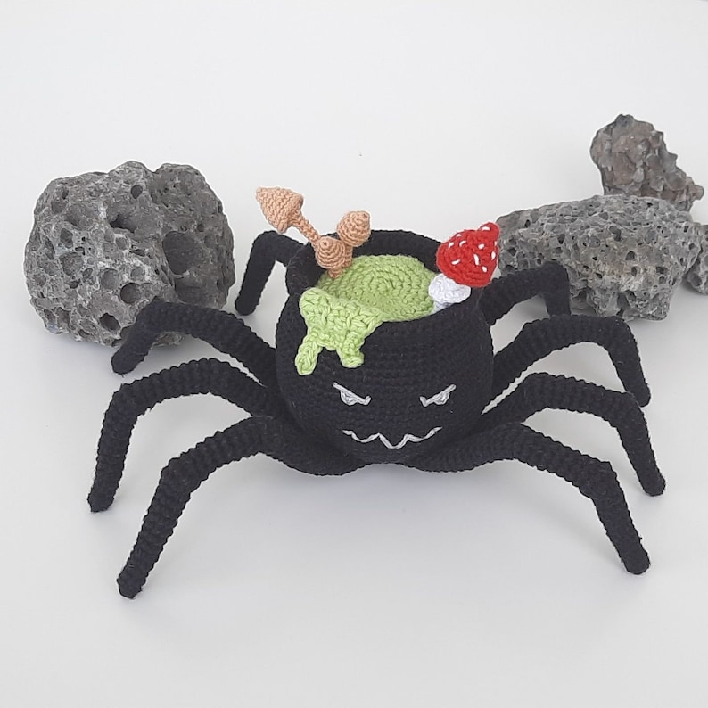 May include: A black crocheted spider with eight legs holding a cauldron filled with green slime and a red mushroom on top.