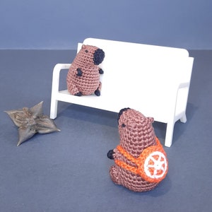 May include: Two brown crocheted capybara figurines sitting on a white bench. One capybara is wearing a small orange backpack with a wheel design.