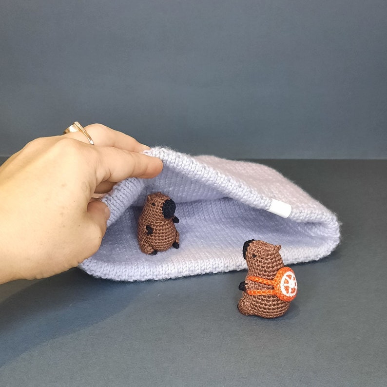 May include: Two crocheted stuffed animals, one brown and one brown with orange accents, are peeking out from a light purple knitted blanket.