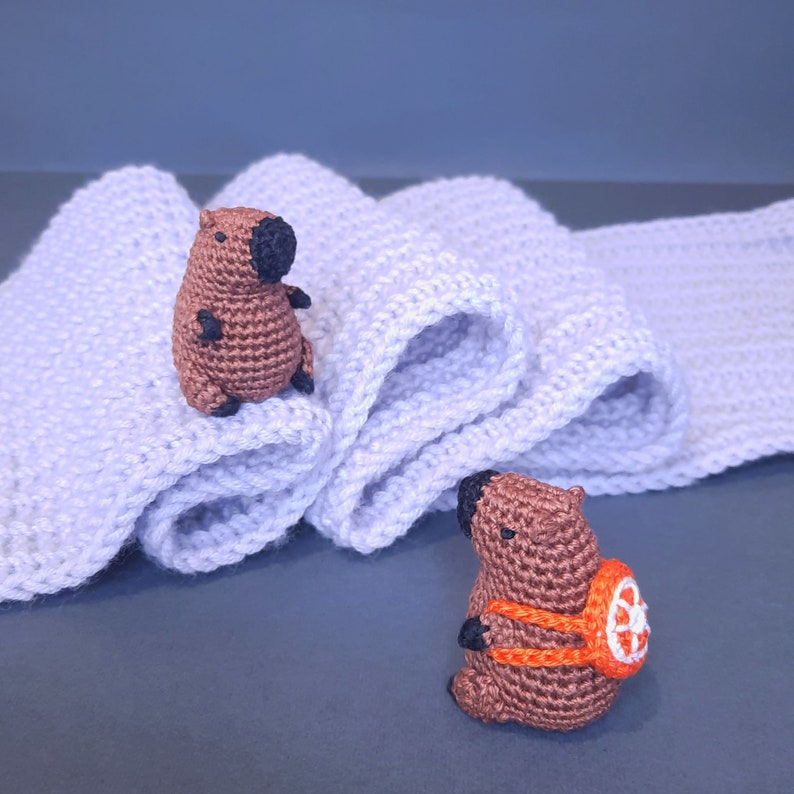 May include: Two crocheted stuffed animals, one brown with black eyes and the other brown with black eyes and an orange backpack, on a light purple crocheted scarf.