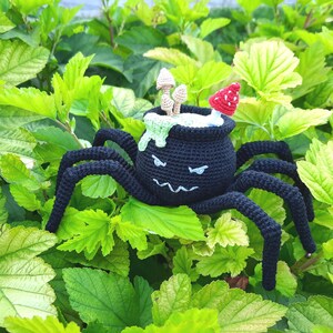 May include: A black crocheted spider with a cauldron on its back. The cauldron is filled with green goo and has a red mushroom on top. The spider is sitting on a bed of green leaves.