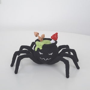 May include: A black crocheted spider with a cauldron on its back. The cauldron is filled with green slime and topped with a red mushroom and brown twigs.