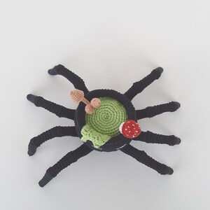 May include: A black crocheted spider with eight legs and a green crocheted pot on its back. The pot has a red mushroom and two brown stems sticking out of it.