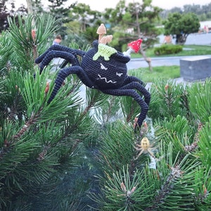 May include: A black crocheted spider with a green cauldron on its back, decorated with mushrooms and a red mushroom cap. The spider is perched on a green pine tree branch.