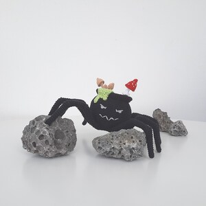 May include: A black crocheted spider with a cauldron on its back. The cauldron has green slime inside and a red mushroom on top. The spider is sitting on a gray rock.