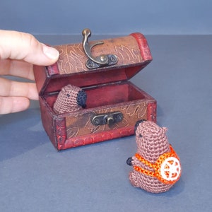 May include: A small wooden treasure chest with a hinged lid and ornate metal accents. The chest is open and contains two small brown crocheted animals.