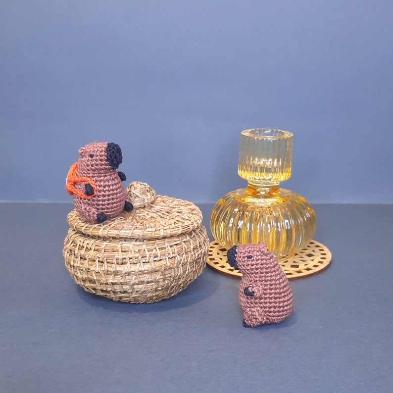 May include: Two crocheted capybara figurines, one sitting on a woven basket with a lid, the other standing on a table. A clear glass vase with a textured surface sits behind the basket.