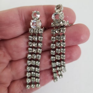 May include: A pair of silver clip-on earrings with a dangling design. Each earring features a cluster of clear rhinestones at the top, followed by a cascade of smaller rhinestones.