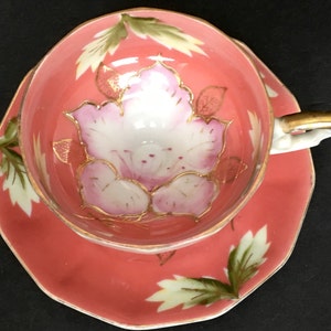 Puede incluir: Un juego de taza y platillo de té floral de color rosa y blanco con detalles dorados. El juego está decorado con una gran flor rosa con ribetes dorados y hojas verdes sobre un fondo coral.