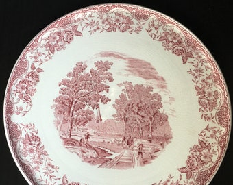 May include: A white ceramic plate with a pink floral border and a pink and white scene of a country road with trees and a church in the background.