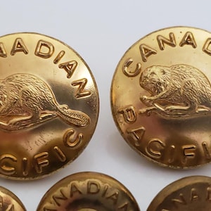 Puede incluir: Primer plano de varios botones dorados. Cada botón presenta un castor en el centro, rodeado de las palabras "CANADIAN PACIFIC". Los botones tienen un aspecto vintage y probablemente sean para ropa o accesorios.