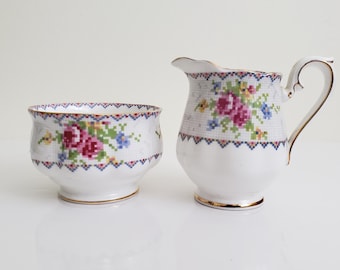 Può includere: Un set di zuccheriera e lattiera in porcellana bianca con un motivo floreale. Il set presenta un delicato disegno floreale con fiori rosa, rossi e blu su sfondo bianco. I bordi della ciotola e della lattiera sono rifiniti in oro.