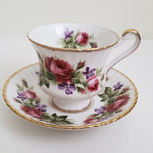 Puede incluir: Una taza de té y un platillo blancos con un patrón floral de rosas rosas y moradas y hojas verdes. La taza y el platillo tienen un borde dorado.