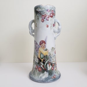 May include: A white porcelain vase with two handles, decorated with a hand-painted scene of a fairy sitting on a branch with berries. The vase has a light blue and green glaze.