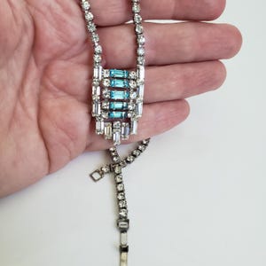 May include: A silver necklace with clear and light blue crystals. The pendant has a rectangular design with rows of clear crystals and a central section of light blue baguette-cut crystals. The necklace has a delicate chain.