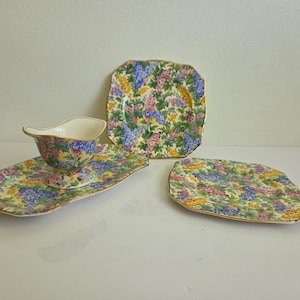 May include: A ceramic set comprising a gravy boat, a long rectangular tray, and two square plates. Each item features a colourful floral pattern with blue, pink, and yellow blossoms on a yellow background, finished with a thin gold trim on the edges.
