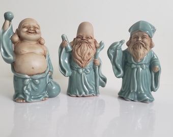 以下が含まれることがあります： 笑顔のローブを着た3体のセラミックフィギュア。フィギュアは青、ベージュ、クリームの色合いです。あるフィギュアはバッグを持ち、別のフィギュアは杖を持ち、3番目のフィギュアは帽子をかぶっています。フィギュアはおそらく装飾品です。