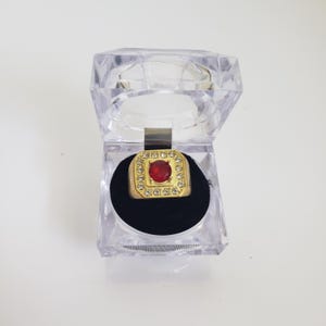 May include: A gold ring featuring a square, red gemstone encircled by smaller clear stones. The ring is presented in a clear, hinged plastic box with a black velvet lining. The ring is a statement piece.