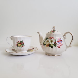 May include: A white porcelain tea set comprising a teapot, teacup, and saucer. The set is adorned with a floral pattern of white flowers, red ribbons, and green leaves. The teacup, saucer, and teapot are edged with gold trim.