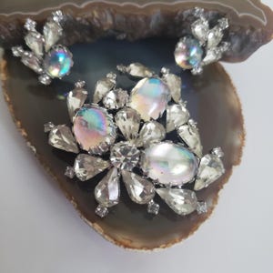 Può includere: Un set di gioielli vintage con spilla e orecchini abbinati. Il set presenta cristalli trasparenti e sfaccettati e pietre iridescenti di forma ovale. La spilla e gli orecchini sono posizionati su una superficie scura e decorata.