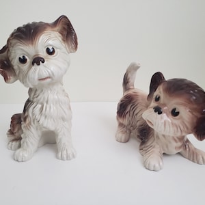 Puede incluir: Dos figuritas de perro de cerámica. El perro de la izquierda está sentado con la cabeza inclinada hacia un lado. El perro de la derecha está de pie con la cabeza girada hacia un lado. Ambos perros son blancos con marcas marrones.