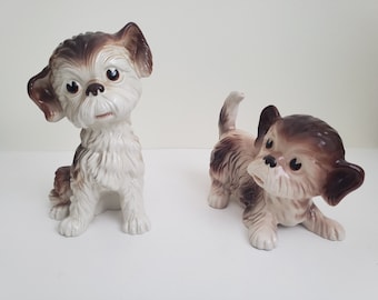 May include: Two ceramic dog figurines. The dog on the left is sitting with its head tilted to the side. The dog on the right is standing with its head turned to the side. Both dogs are white with brown markings.
