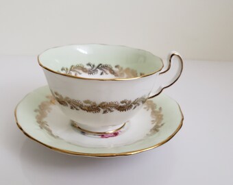 May include: A white teacup and saucer with a green and gold floral pattern. The cup has a gold rim and the saucer has a scalloped edge.