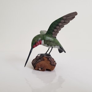 Op de afbeelding: Houten beeldhouwwerk van een kolibrie met uitgespreide vleugels, geplaatst op een kleine, bruine, getextureerde basis. De vogel heeft groene en bruine vleugels, een rode keel en een lange, zwarte snavel. De sculptuur staat tegen een witte achtergrond.