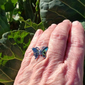 May include: A silver butterfly ring with iridescent blue and green wings. The ring is a close-up shot, showcasing the intricate details of the butterfly design. The ring is on a person's hand, with a blurred green leafy background.