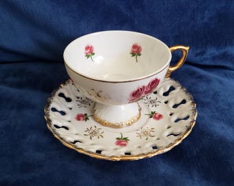 Puede incluir: Juego de taza y platillo blancos con diseño de rosas. La taza tiene un asa dorada y está sobre una base de pedestal. El platillo tiene un diseño calado decorativo con ribete dorado.