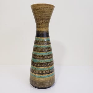 May include: A tall, brown ceramic vase with a geometric pattern in shades of blue, green, and brown. The vase has a narrow neck and a wide base.