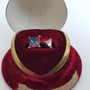 May include: Two rings with gold bands and gemstone settings are presented in a heart-shaped, red velvet jewellery box. One ring has a square, iridescent gemstone, the other a triangular, clear gemstone.