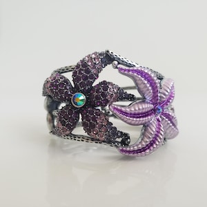 May include: A silver-toned cuff bracelet featuring two flower designs. One flower is covered in small, dark purple crystals, and the other is a light purple starfish shape. Both flowers have a central iridescent crystal.
