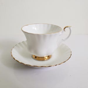 May include: A white teacup and saucer with gold detailing. The teacup has a fluted design and a gold base. The saucer has a scalloped edge, also with gold trim. A classic tea set, perfect for afternoon tea.
