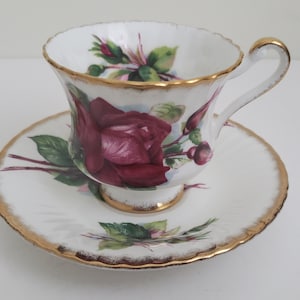 May include: A white teacup and saucer with a gold rim and a floral design featuring a large pink rose and green leaves.