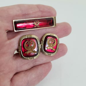 May include: A vintage set of cufflinks and a tie clip. The set includes a rectangular tie clip and two square cufflinks. They have a red background and gold-coloured trim. Each piece features a gold emblem with a star, crescent, and sword design.