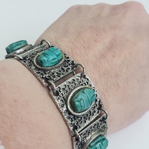 Op de afbeelding: Zilverkleurige armband met turkoois scarabee-ontwerpen. De armband heeft een opengewerkt filigraanpatroon en ovale turkooizen stenen. De armband is verbonden door kleine zilveren ringen.