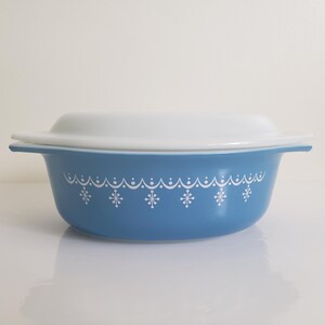 May include: A blue casserole dish with a white lid. The dish has a white snowflake and scalloped design around the rim. The dish has two handles and is sitting on a white surface.