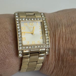 May include: A gold-tone rectangular wristwatch with a white face and gold-tone numerals. The bezel is encrusted with clear crystals. The watch features a gold-tone metal link bracelet and gold-tone hands. The word GUESS is printed on the watch face.