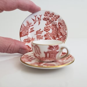 Op de afbeelding: Een miniatuur theeset met een kopje, schotel en een klein bord. De set heeft een witte achtergrond met een rood en wit Willow patroon ontwerp. De randen van de schotel en het bord zijn afgewerkt met goud. De set wordt door een hand vastgehouden.