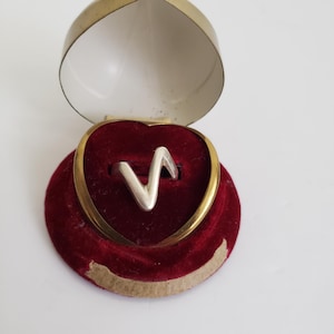 Peut inclure: Une bague argentée avec un motif en zigzag est présentée dans un écrin en forme de cœur. L'écrin a un intérieur en velours rouge et un bord doré. L'écrin est ouvert, révélant la bague. L'extérieur de l'écrin est doré avec un intérieur blanc.