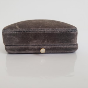 May include: A small, rectangular jewellery box with a rounded top. The box is covered in a soft, grey velvet material. A small, round, off-white button serves as a clasp on the front. The box is designed to hold jewellery.