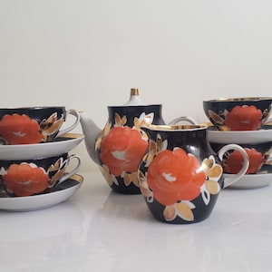 May include: A vintage tea set featuring a teapot, creamer, sugar bowl, and six teacups with saucers. The set is navy blue with large orange floral designs and gold trim. The teacups and saucers are stacked in pairs.