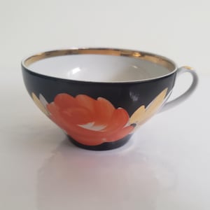 May include: A black teacup with a gold rim and a white interior. The cup features a large orange and yellow floral design on the exterior and a white handle.