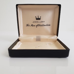 Puede incluir: Una caja de joyería negra abierta con un interior color crema. La caja tiene las palabras "JEWELLERY" y "For Men of Distinction" impresas en el interior. Un pequeño gráfico de corona está encima de la palabra "JEWELLERY".