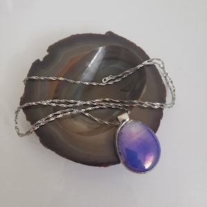 Puede incluir: Un collar de cadena de plata con un gran colgante ovalado de vidrio iridiscente. El colgante es de color morado oscuro con toques de rosa y azul.