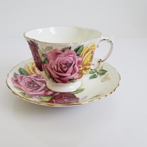 Puede incluir: Un juego de taza y platillo de té vintage. La taza y el platillo están decorados con rosas rosas y amarillas y hojas verdes. Los bordes están adornados con oro. La taza tiene un asa y se apoya sobre un pie pequeño.