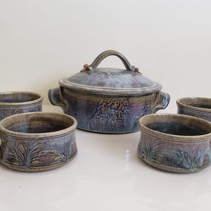 Può includere: Un set di quattro ciotole per zuppa in ceramica marrone e blu con una pentola per zuppa in ceramica abbinata con coperchio. Le ciotole e la pentola hanno un aspetto rustico e artigianale con un design di piante stilizzate.
